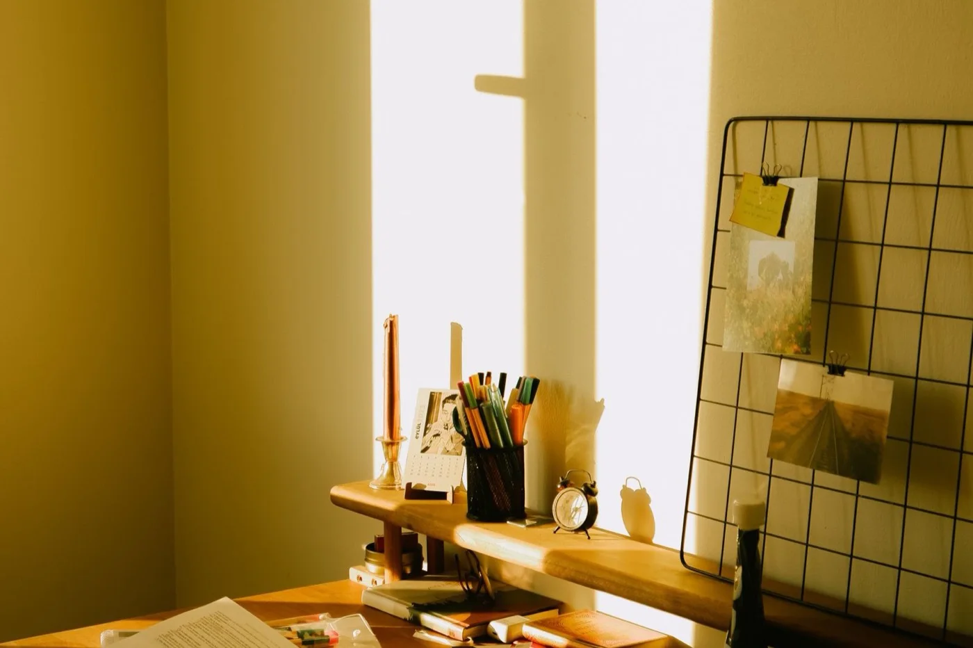 Schreibtisch vor einem Fenster, Sonnenlicht auf gelber Wand, Stiftbecher, Notizen und ein aufgeschlagenes Buch