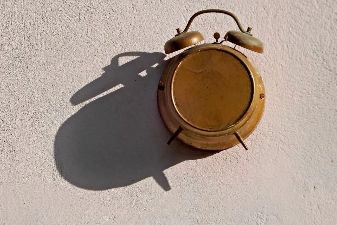 Messingfarbener Wecker an einer hellen Wand, sein Schatten fällt deutlich größer daneben