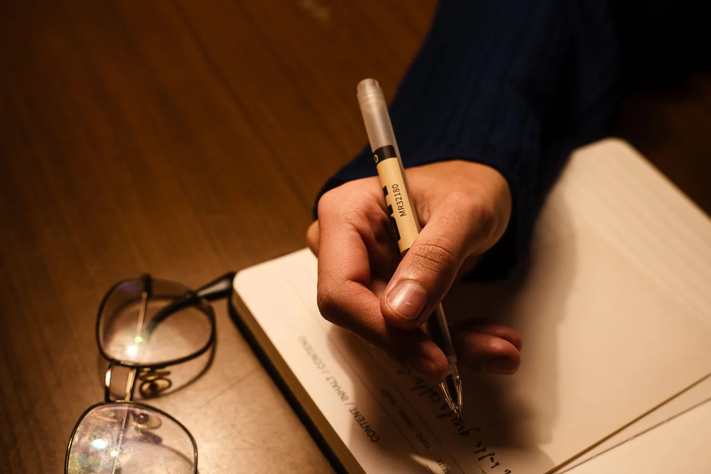 Hand mit Stift schreibt bei warmem Lampenlicht in ein aufgeschlagenes Notizbuch, daneben eine Brille auf dunklem Holztisch