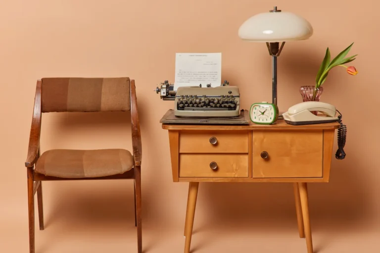 Vintage-Arbeitsplatz vor terrakottafarbener Wand: Holzstuhl, Schreibtisch mit Schreibmaschine, Lampe, Wecker und Telefon